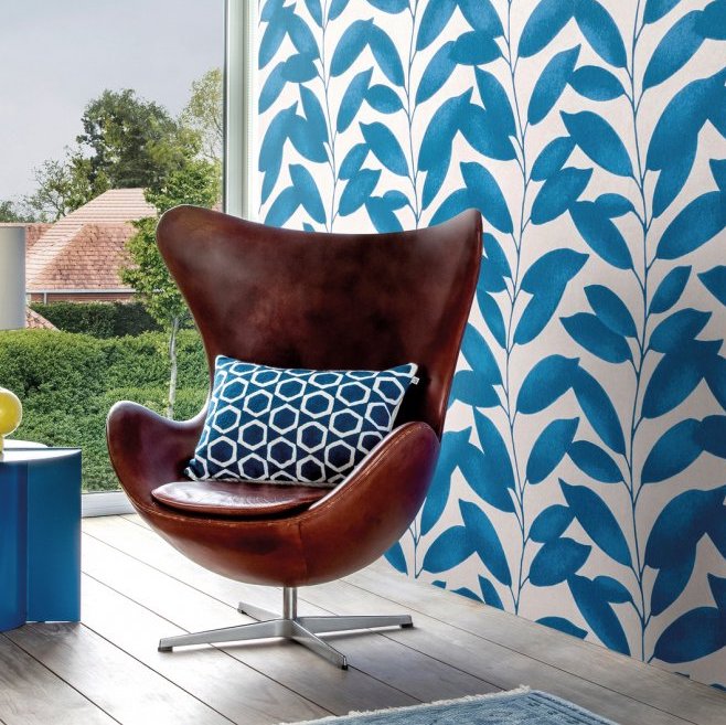 Bruine designstoel met blauw sierkussen voor een muur met blauw-wit Arte-behang uit de Joy Blooming Marvellous-collectie.