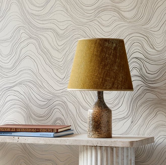 Tafel met gouden lamp en boeken tegen Arte-behang Kharga Bayuda 74042 met golvend lijnpatroon.