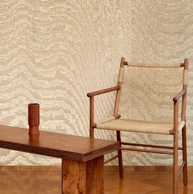 Stoel van hout en touw staat naast een houten tafel met decoratief object, tegen Arte-behang Kharga Moiré 15011A in bright gold.