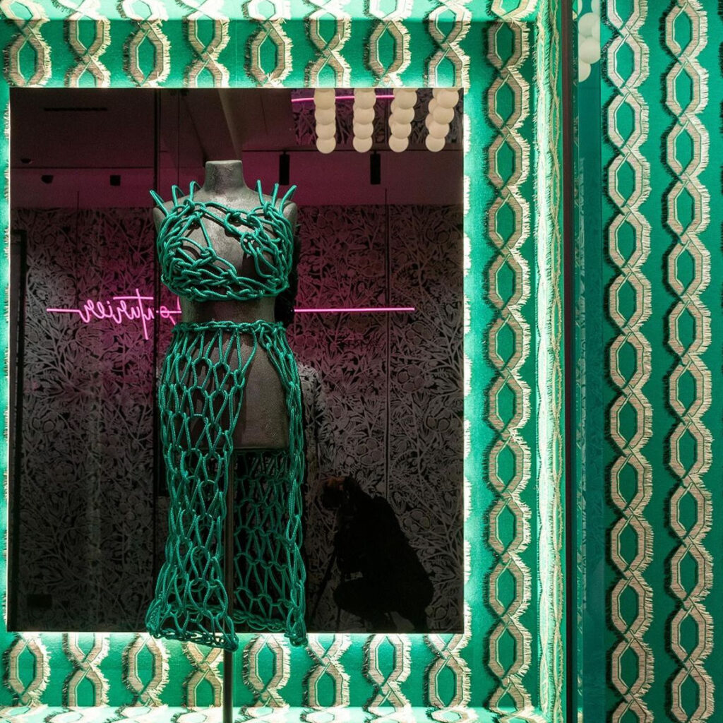Etalage met een kunstzinnig groen macramé-jurk op een paspop, omlijst door het opvallende groene Arte-behang Le Couturier Franges 58520.
