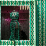 Etalage met een kunstzinnig groen macramé-jurk op een paspop, omlijst door het opvallende groene Arte-behang Le Couturier Franges 58520.