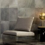 Designstoel met metalen frame en zacht kussen staat voor een muur bekleed met Arte-behang Les Cuirs Rectangle.