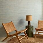 Twee rotan stoelen en een lamp op een groene bijzettafel staan op een juten vloerkleed tegen een wand met Arte Ruban Palmier behang.