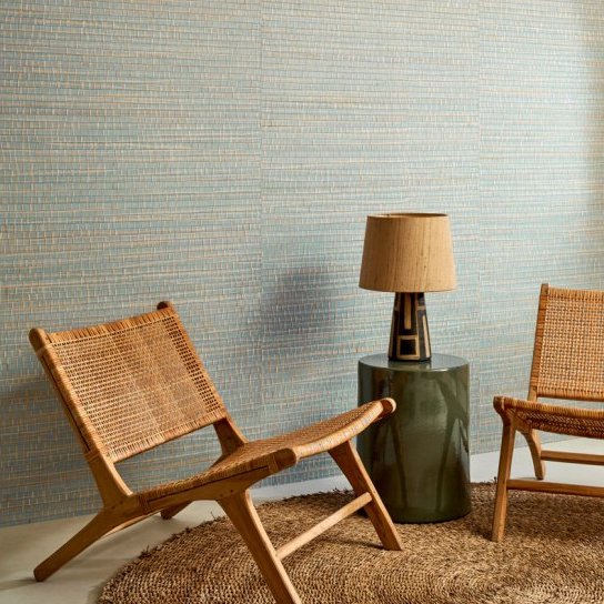 Twee rotan stoelen en een lamp op een groene bijzettafel staan op een juten vloerkleed tegen een wand met Arte Ruban Palmier behang.