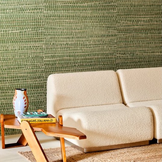 Crèmekleurige bank en houten salontafel met vaas tegen een groene Arte Ruban Palmier Les Forêts behangmuur.