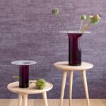 Twee paarse vazen met bloemen staan op houten bijzettafels voor een paars Arte Textura Canvas behang.