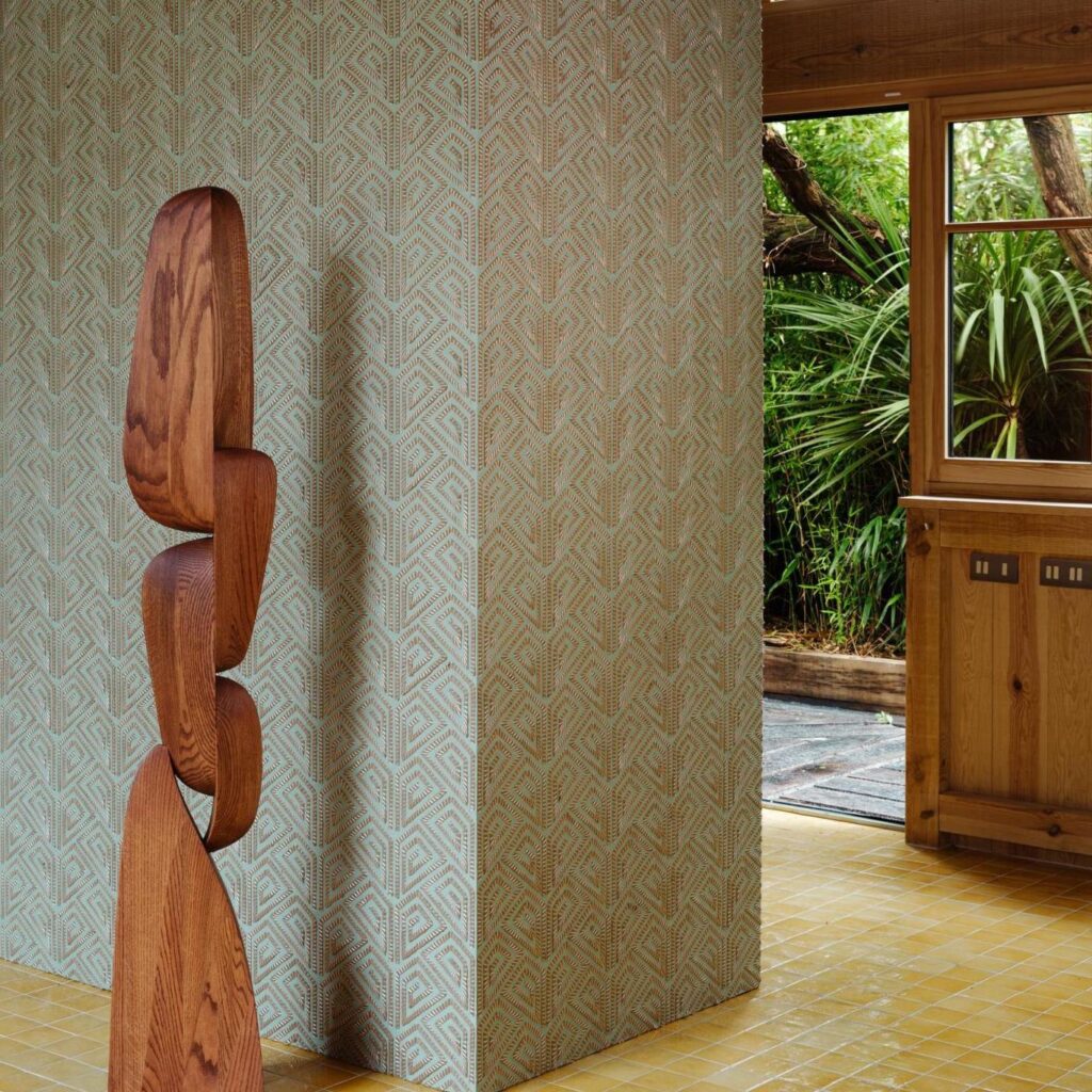 Houten sculptuur voor een met Arte Carus Alata 15712 behangen muur in een interieur met zicht op een tuin.