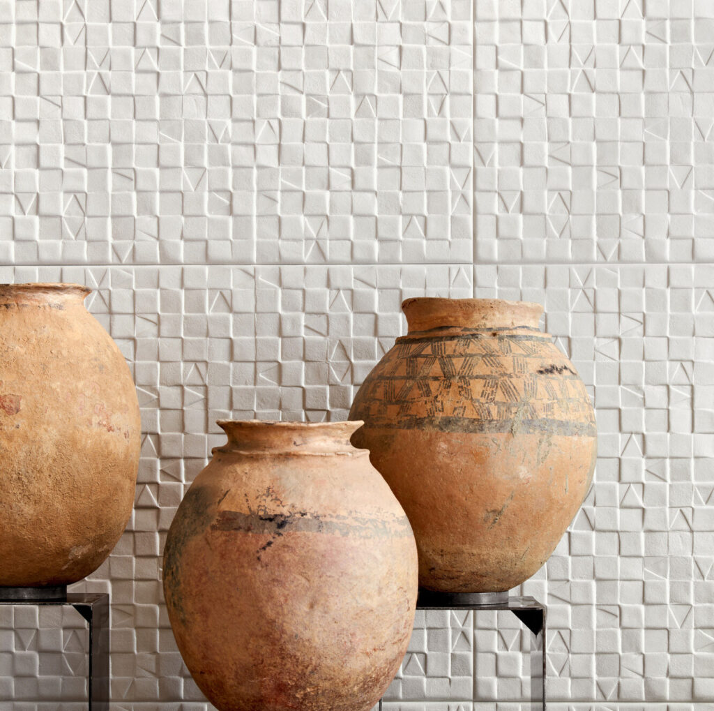 Drie aardewerken vazen staan voor een witte Babylon Adobe wand met een geometrisch reliëfpatroon.