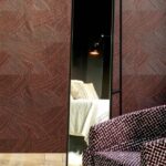 Kamer met Arte behang Arado Rose Brown, een spiegel die een bed reflecteert, en een stoel met grafisch patroon op de voorgrond.