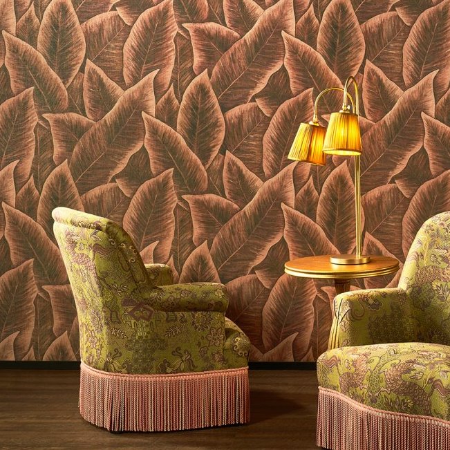 Twee klassieke fauteuils en een bijzettafel met lamp staan voor behang met tropisch bladerpatroon uit de Arte Bird of Paradise-collectie.
