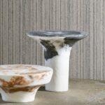 Twee organisch gevormde bijzettafels van marmerachtige materialen voor een muur met Arte Linea behang met verticale textuur.