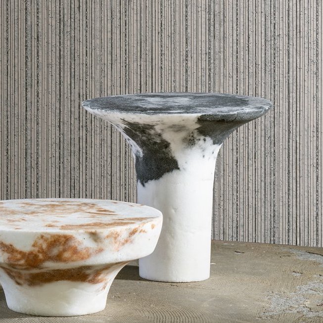 Twee organisch gevormde bijzettafels van marmerachtige materialen voor een muur met Arte Linea behang met verticale textuur.