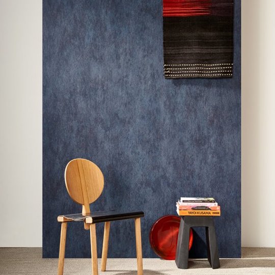 Moderne interieursetting met Arte Manchas behang in blauw, een houten designstoel, een zwarte bijzettafel met boeken en een rood decoratief object.