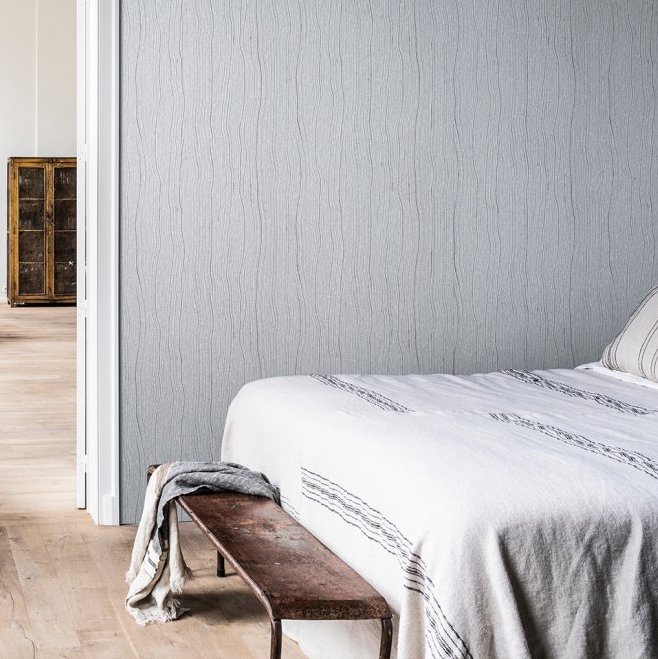Slaapkamer met Arte Timber behang in een subtiel houtnerfpatroon, naast een bed met witte linnen en houten bankje.