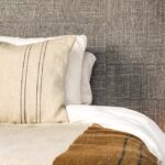 Close-up van een bed met witte en beige lakens en kussens, voor een muur met Arte behang uit de Twine collectie.