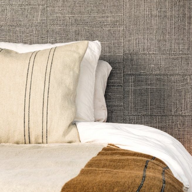 Close-up van een bed met witte en beige lakens en kussens, voor een muur met Arte behang uit de Twine collectie.