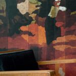 Detail van het Elitis Cordoba VP 926 01 behang met abstract bloemmotief in warme tinten achter een houten stoel met zwarte bekleding.