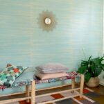 Interieur met Elitis behang Kari VP 930 40 in turquoise tint, een houten bank met kussens, een groene plant en een decoratieve spiegel aan de muur.
