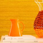 Oranje kan en decoratief object op boeken voor Elitis Kosa Silk behang in een warme, goudgele tint.