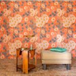 Interieur met Elitis Margaritas behang in oranje bloemmotief, een houten bijzettafel met lamp en een beige poef met groen kussen.