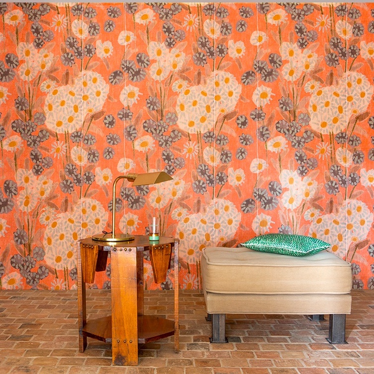 Interieur met Elitis Margaritas behang in oranje bloemmotief, een houten bijzettafel met lamp en een beige poef met groen kussen.