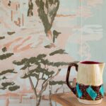 Decoratief Elitis Mediterranée behang met bomen en rotsformaties in zachte pastelkleuren, met een keramische kan op een houten oppervlak op de voorgrond.