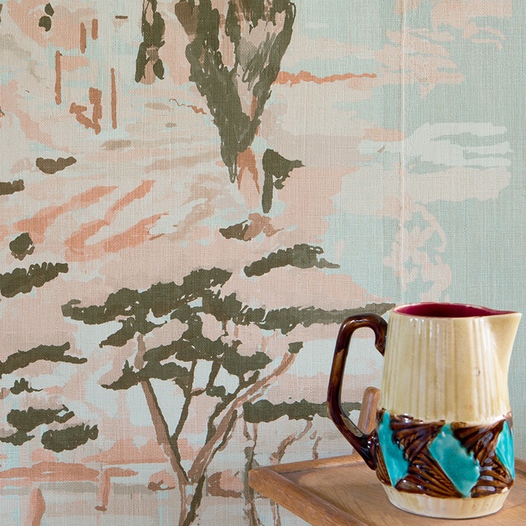 Decoratief Elitis Mediterranée behang met bomen en rotsformaties in zachte pastelkleuren, met een keramische kan op een houten oppervlak op de voorgrond.