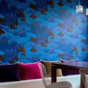 Interieur met het blauwe Elitis behang 'Rêves de Manille TP 323 05', kleurrijke fluwelen kussens en een houten tafel met moderne stoelen.