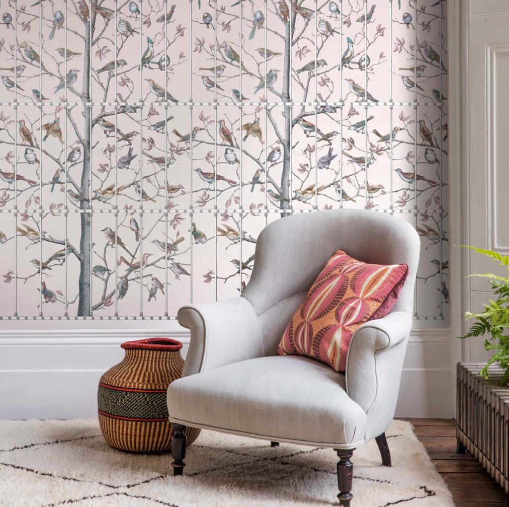 Grijze fauteuil met sierkussen voor Fornasetti Uccelli-behang met vogel- en boommotief.