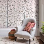 Grijze fauteuil met sierkussen voor Fornasetti Uccelli-behang met vogel- en boommotief.