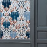 Decoratief Lelievre behang (model CAP D'AIL 6470-03) met symmetrisch patroon van bladeren en gestileerde gezichten in blauw, roodbruin en wit.
