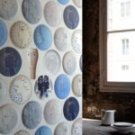 Behang met abstracte, keramiekachtige gezichten in blauwe, grijze en neutrale tinten uit de collectie POTERIES, naast een rustiek raam.