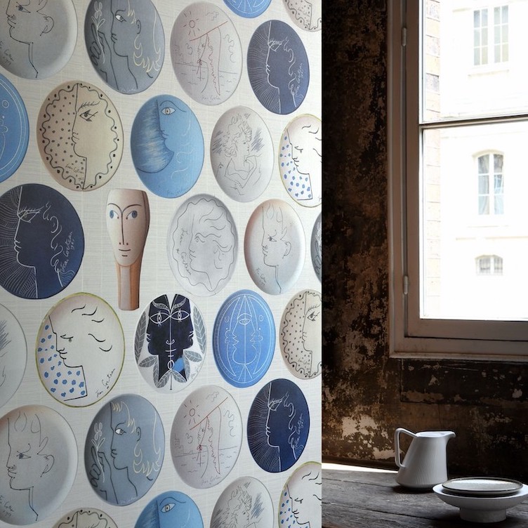 Behang met abstracte, keramiekachtige gezichten in blauwe, grijze en neutrale tinten uit de collectie POTERIES, naast een rustiek raam.