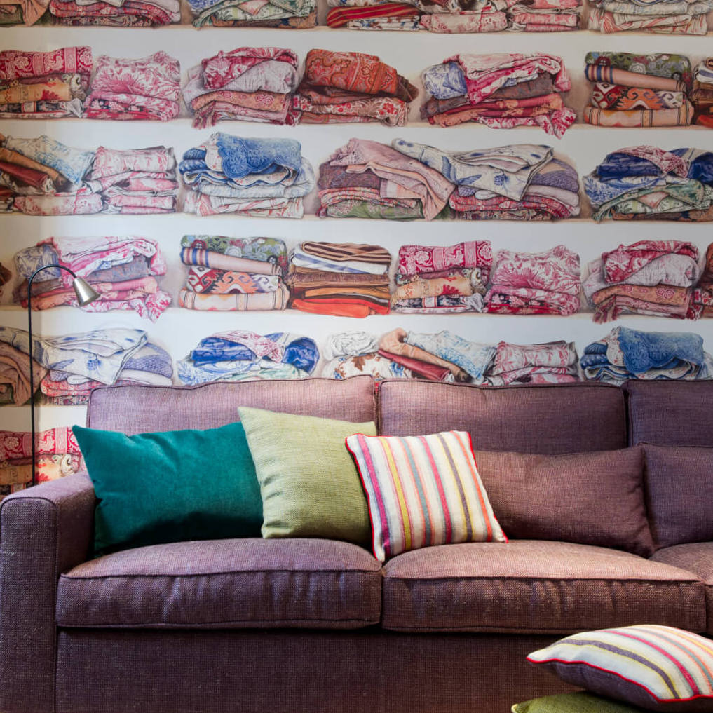 Paarse bank met gekleurde kussens voor Pierre Frey behang FP319001 met een motief van opgevouwen dekens.