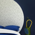 Slaapkamer met opvallend RESSEMBLANCE-behang in een grote cirkelvorm op een donkerblauwe muur, gecombineerd met blauw beddengoed en een groene vaas met decoratieve objecten.