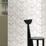 Wit behang met een herhalend abstract lijnpatroon van tegenovergestelde gezichten, genaamd TÊTE À TÊTE, met een zwarte designstoel op de voorgrond.
