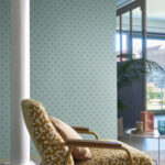 Stijlvol interieur met CASAMANCE Apis-behang in lichtblauw met subtiel patroon, naast een retro fauteuil en grote ramen.