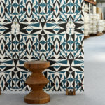 Geometrisch behang met abstracte gezichten en blauw-zwarte patronen in een industriële ruimte, met een houten kruk ervoor.