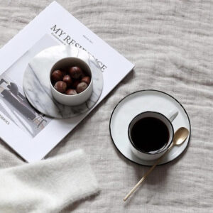 Witte kop koffie met schotel en goudkleurige lepel op een Broste Copenhagen tafelkleed in de kleur Simply Taupe, naast een tijdschrift en een schaaltje met noten.