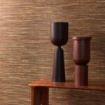 Twee houten decoratieve objecten op een houten tafel voor Buningh Arte Geloma behang met een warm, geweven structuurpatroon.