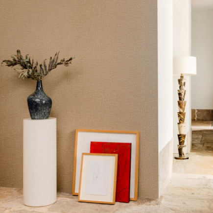 Interieurhoek met een beige Lin Brodés wandbekleding uit de Buningh Elitis collectie, gedecoreerd met een vaas op een witte sokkel en ingelijste kunstwerken op de vloer.