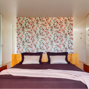 Slaapkamer met Buningh Elitis Milos behang met bloesemmotief achter een houten bed met paarse en grijze accenten.