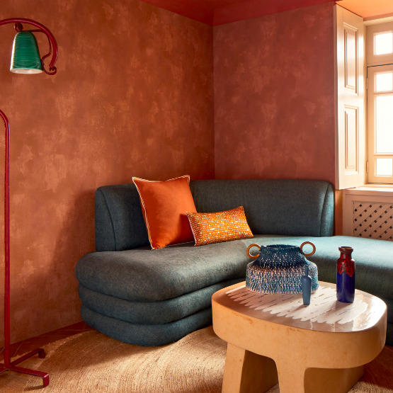 Sfeervol ingerichte woonkamer met Buningh-Elitis Ortigia Agrigente behang, een blauwe designbank en terracotta accenten.