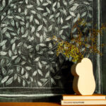 Witte organische vaas met takjes op een stapel boeken voor een muur met het Buningh-Elitis Ortigia Disegno VP 961 01 bladerenpatroon.