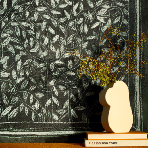 Witte organische vaas met takjes op een stapel boeken voor een muur met het Buningh-Elitis Ortigia Disegno VP 961 01 bladerenpatroon.