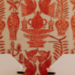 Behang uit de Buningh-Elitis Paros collectie met rood-oranje geborduurde motieven van vogels, vazen en planten op een beige achtergrond.