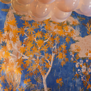 Detail van het Buningh Elitis Retour des Indes behang uit de Ortigia collectie met goudkleurige bladeren op een blauwe achtergrond en een lichtarmatuur van glasbollen erboven.