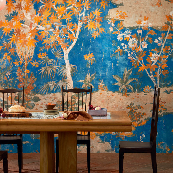 Eetkamer met houten tafel en stoelen tegen een kleurrijk behang uit de Ortigia-collectie met oriëntaalse bomen en planten op een blauwe achtergrond.