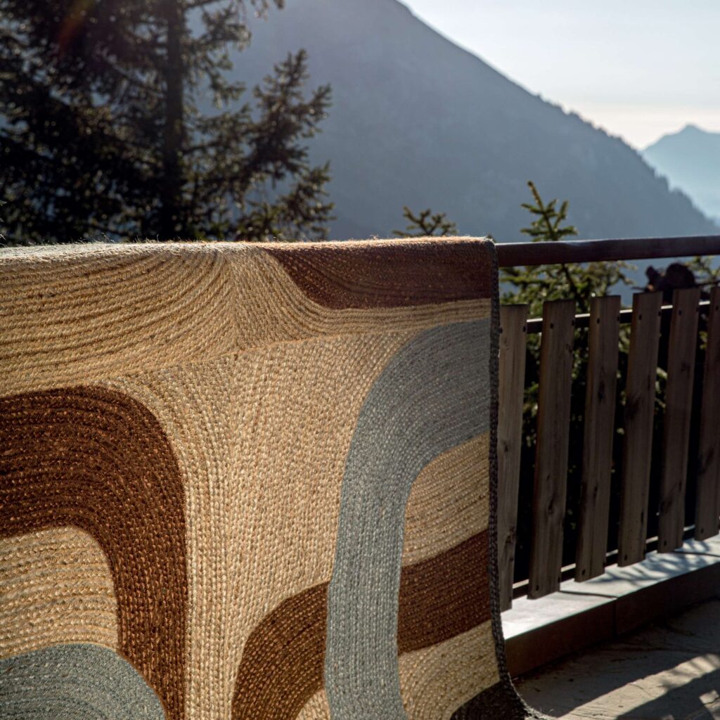 Vloerkleed Elitis Penny Lane TA 11942 in bruin- en blauwtinten hangt over een houten balkonreling met uitzicht op berglandschap.
