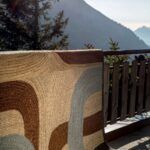 Vloerkleed Elitis Penny Lane TA 11942 in bruin- en blauwtinten hangt over een houten balkonreling met uitzicht op berglandschap.